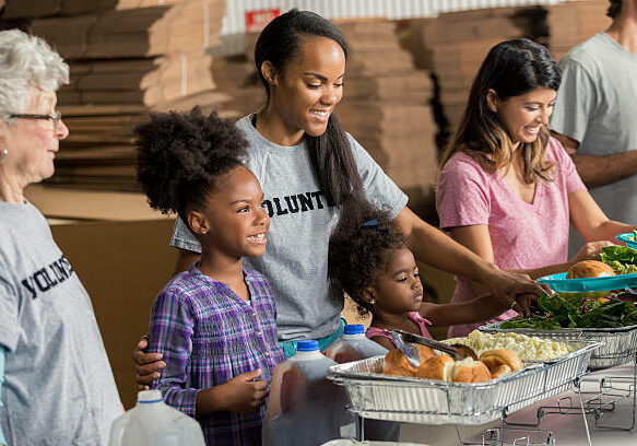 African American mother and her two daughters serve meals to the homeless with their diverse family.